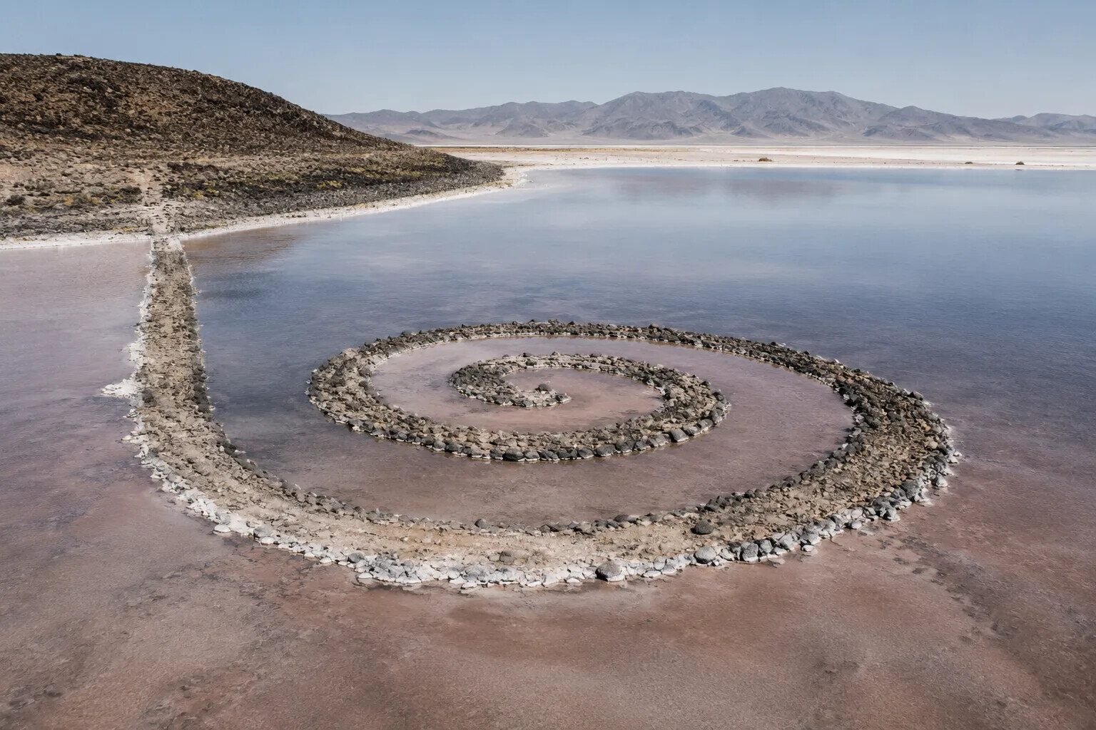 Aerial view of a spiral stone jetty extending into a shallow lake, inspired by Robert Smithson's Spiral Jetty and illustrating the Land Art movement.
